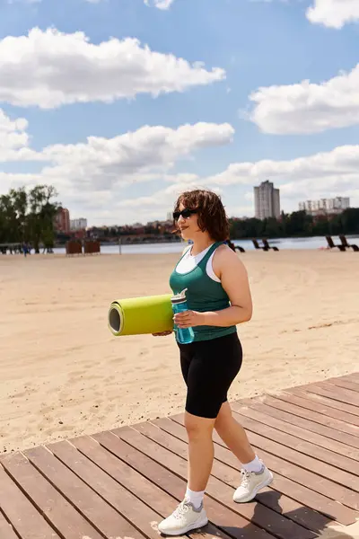 A sporty woman with a yoga mat walks along the waterfront enjoying the lazy afternoon sun. — Stock Photo