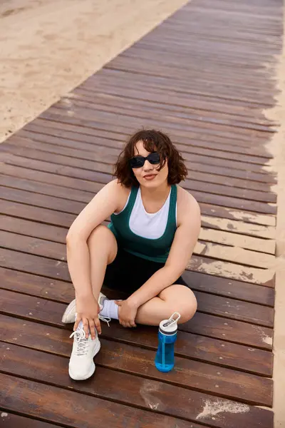 Una joven descansa en un camino de madera después de un entrenamiento en la playa, mostrando su forma física. - foto de stock
