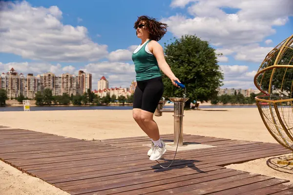 A fit woman jumps rope outdoors by the river, surrounded by city buildings on a sunny day. — Stock Photo
