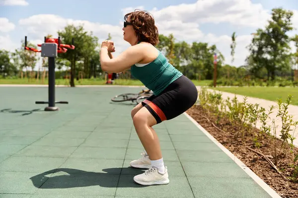 A young and fit woman exercises outdoors, engaging in squats amid lush greenery and clear skies. — Stock Photo