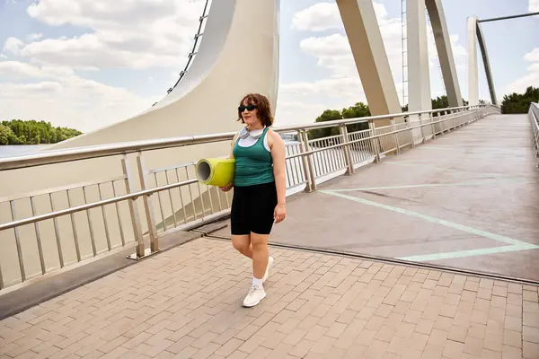 A young woman strides confidently along a bridge, carrying exercise gear under a clear sky. — Stock Photo