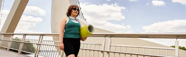 Una donna giovane e in forma fa una passeggiata su un ponte mentre porta un tappetino da yoga, striscione — Foto stock