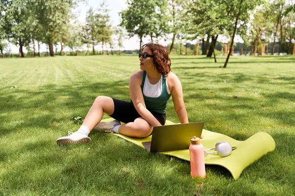 Una joven se relaja en un día soleado, trabajando en su portátil en la hierba. - foto de stock