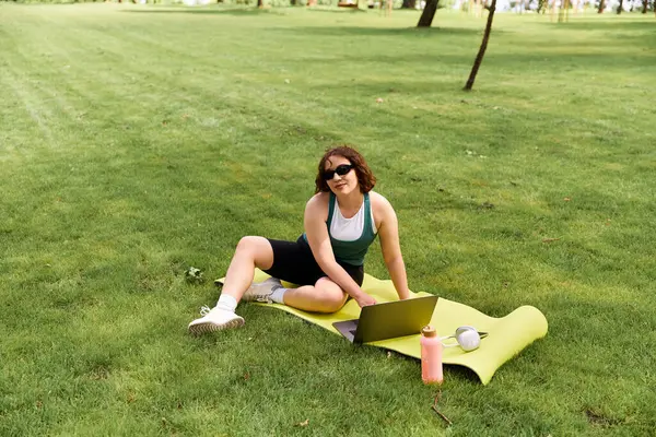 Una giovane e graziosa donna si rilassa su un tappetino yoga, lavorando sul suo computer portatile sotto il sole estivo. — Foto stock