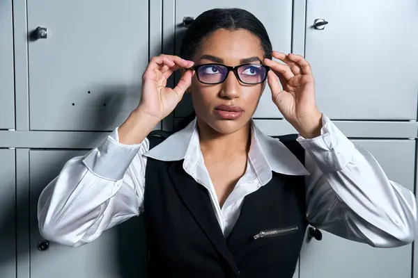 Mujer joven ajusta sus gafas con estilo mientras exuda elegancia en un traje de moda en medio de casilleros. - foto de stock