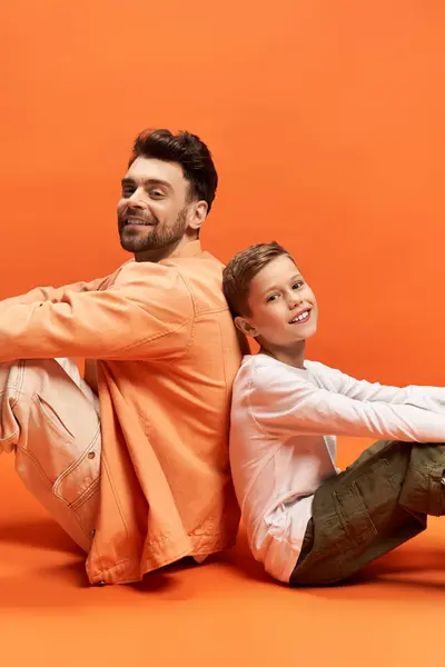 A father and son share a heartwarming moment, smiling together against a vibrant orange background. — Stock Photo