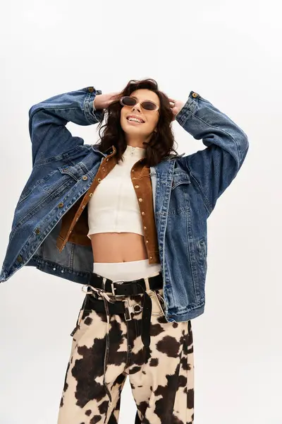 Trendy young woman poses joyfully in cow print pants and denim jacket against a grey backdrop. — Stock Photo