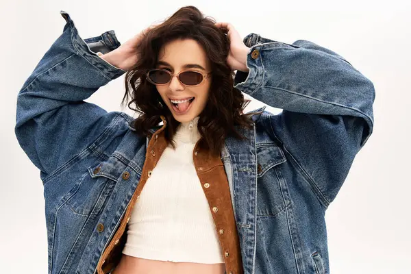 A fashionable young woman poses in chic sunglasses and denim jacket and sticking out tongue against a neutral backdrop. — Stock Photo