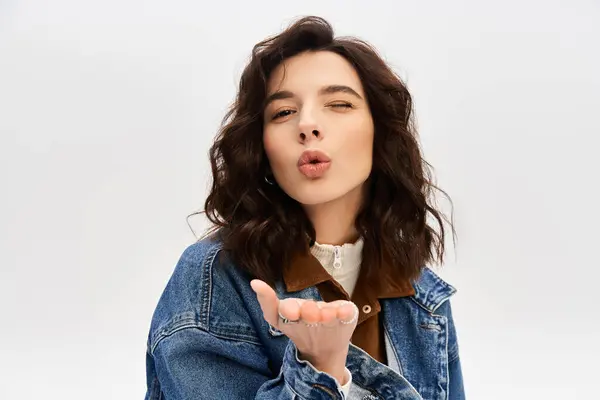 A stylish young woman blows a kiss in trendy outfit against a grey backdrop. — Stock Photo