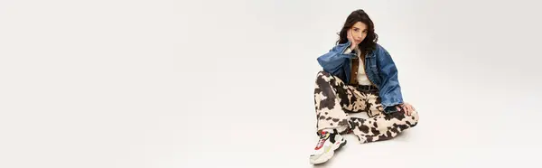 A young woman sits elegantly wearing cow print pants and a denim jacket against a grey backdrop, banner — Stock Photo