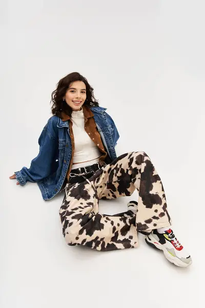 A young woman wearing cow print pants and a denim jacket poses playfully against a grey backdrop. — Stock Photo
