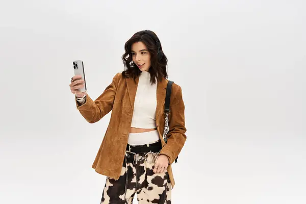 Young woman poses confidently in a fashionable outfit against a grey backdrop while taking a selfie. — Stock Photo