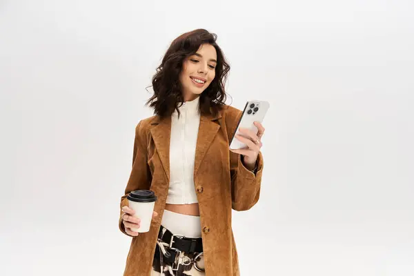 Charming young woman smiles while checking her phone, holding a coffee cup in her trendy outfit. — Stock Photo