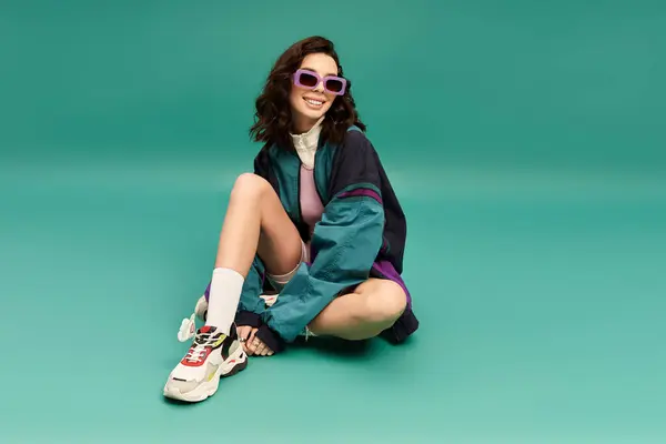 Sporty young woman in a trendy outfit relaxes confidently against a vibrant turquoise backdrop. — Stock Photo