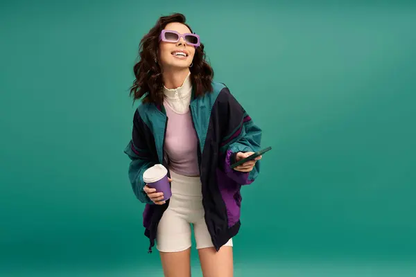 A cheerful young woman in trendy sportswear stands confidently, holding a drink and phone. — Stock Photo