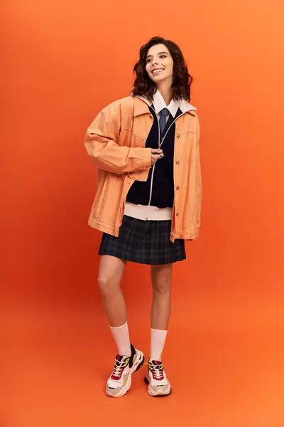 Young woman poses with confidence in fashionable skirt and jacket on bright orange backdrop. — Stock Photo