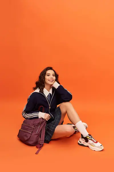 A young woman in a trendy outfit sits gracefully against an orange backdrop, exuding confidence. — Stock Photo