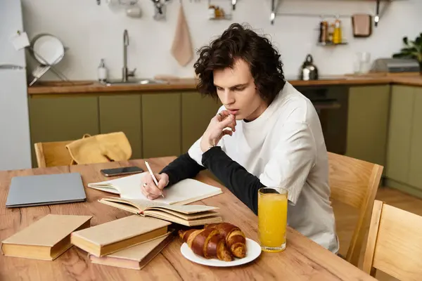 Young man sits at a wooden table, writing in a journal, savoring breakfast with orange juice. — Photo de stock