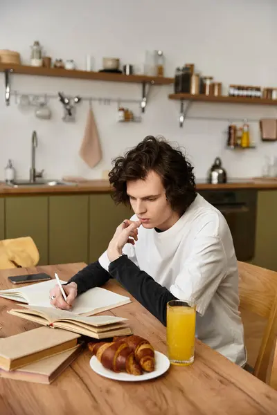 A young man sits at a wooden table, writing in a notebook while enjoying croissants and juice. - foto de stock