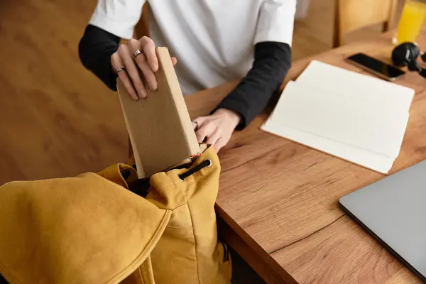 A relaxed young man in cozy attire unpacks a book from his yellow bag at home. — стокове фото