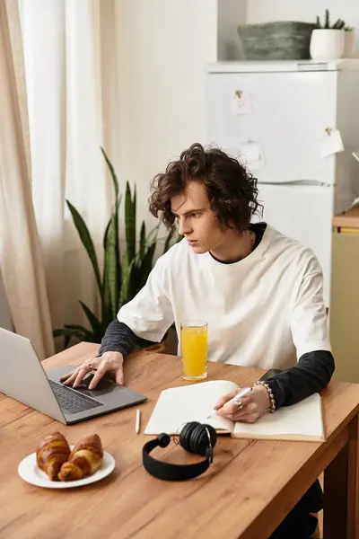 A young man sits at home, writing in a notebook and enjoying a meal while using his laptop. — Stockfoto