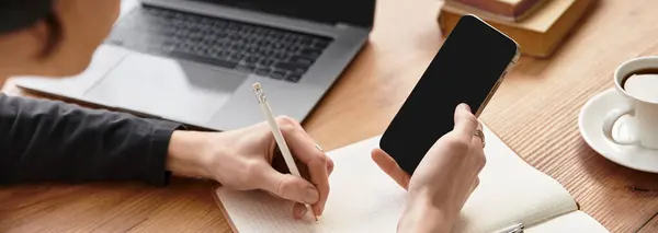 A young man in cozy attire focuses on jotting down ideas while using his smartphone at home. - foto de stock