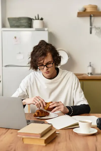 A young man in comfy clothes enjoys a pastry while working on his laptop at a wooden table. — Stock Photo