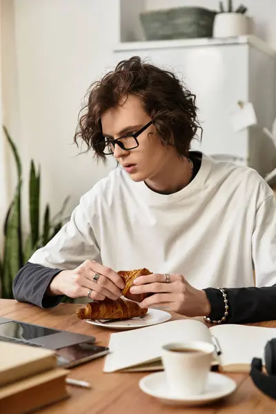 A young man with curly hair enjoys a croissant while focused on his laptop at home. — Photo de stock