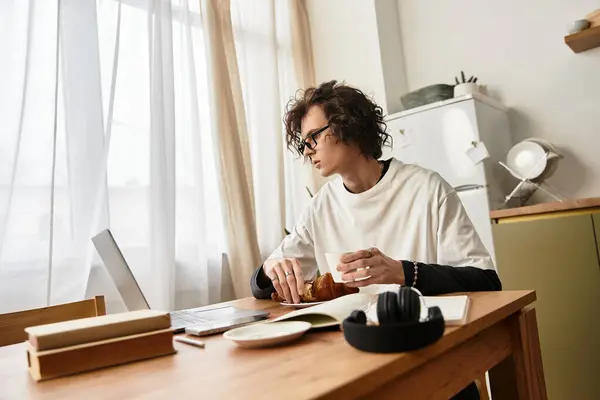 A young man in casual attire sits at a wooden table, focused on his laptop while sipping a drink. — Photo de stock