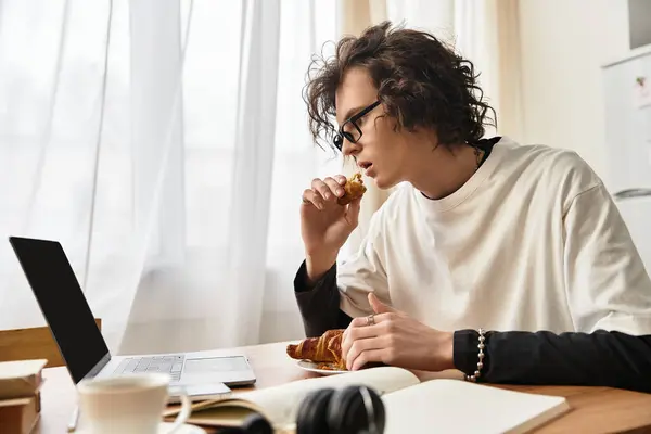 A young man bites into a pastry while seated at a wooden table, using a laptop to connect online. — Stockfoto