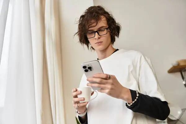 Ein junger Mann entspannt sich in bequemen Klamotten, nippt am Kaffee und blättert in seinem Handy drinnen. — Stockfoto