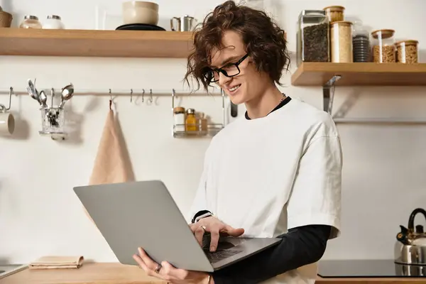 A handsome young man in comfortable attire focuses on his laptop in a warm kitchen atmosphere. — Stock Photo