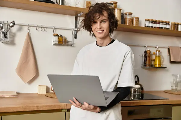 Smiling young man in casual attire stands in his warm kitchen, engaged with his laptop. — Stock Photo
