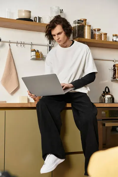 A young man in cozy clothes sits on his kitchen counter, focused on his laptop. — Stock Photo