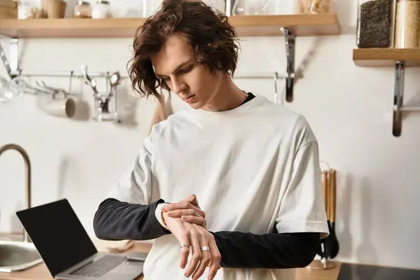A young man in relaxed attire glances at his watch while seated in a modern kitchen. - foto de stock