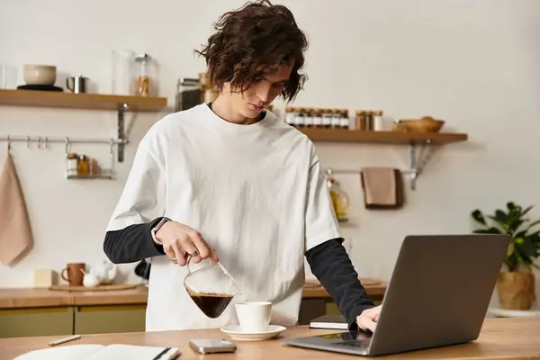 A young man in comfortable attire pours coffee into a cup while focused on his laptop. — Photo de stock