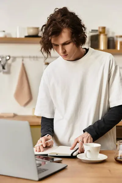 A handsome young man wearing relaxed attire writes notes at his kitchen table while sipping coffee. — Stock Photo