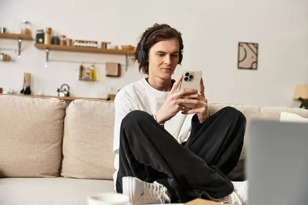 Um jovem senta-se confortavelmente em roupas aconchegantes, usando seu telefone para ouvir música em casa. — Fotografia de Stock