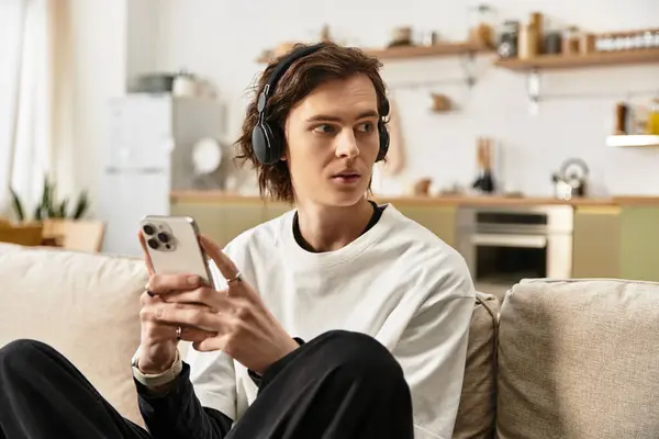A young man relaxes on a couch, focused on his smartphone with stylish headphones on. — Stock Photo