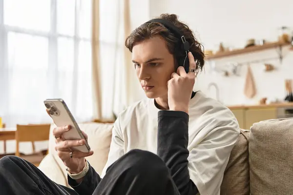 A young man sits comfortably on a couch, listening to music with headphones while using his phone. — Stock Photo