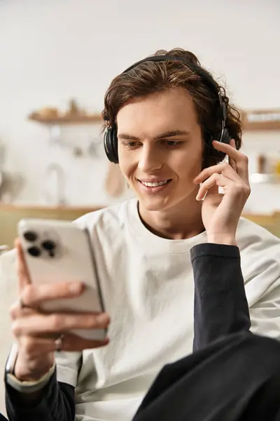 In a comfortable home setting, a young man listens to music with headphones while smiling. - foto de stock