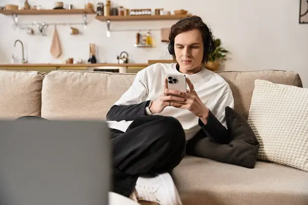 Young man sits on a soft couch at home, focused on his smartphone while in cozy clothing. — Stock Photo