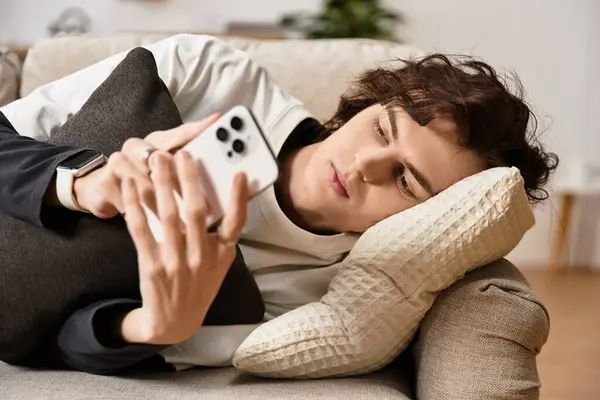 A young man comfortably lounges on a sofa, absorbed in his smartphone, wearing casual clothing. — Fotografia de Stock