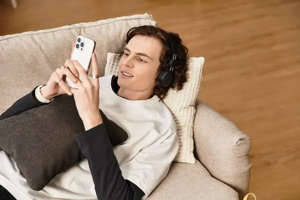 A young man lounges comfortably on a sofa, engaging with his smartphone, dressed casually. — Fotografia de Stock