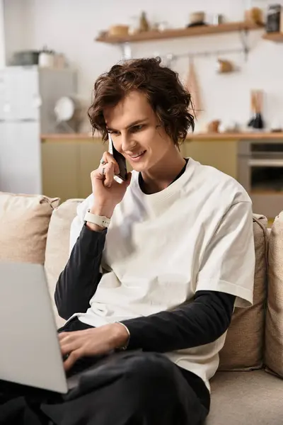 A young man in comfy clothes smiles on a phone call while multitasking on his laptop. — Stock Photo