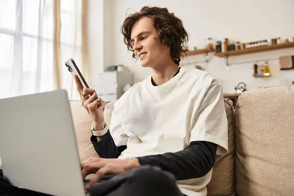 A young man dressed in cozy attire relaxes at home, focused on his smartphone and laptop. — Foto stock