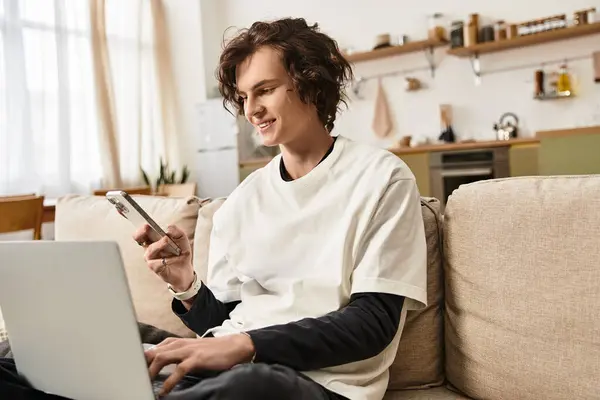 A young man dressed comfortably sits on a sofa, using a laptop and smartphone, smiling with joy. — Fotografia de Stock