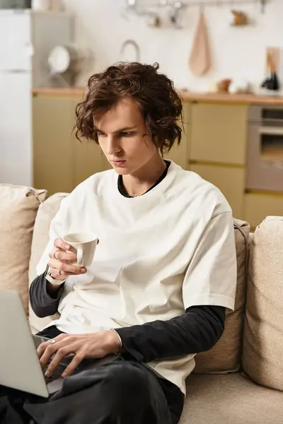 Relaxed young man in a cozy outfit focuses on his laptop while sipping coffee at home. — Photo de stock