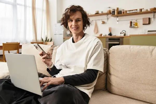 A young man relaxes at home, wearing cozy attire, while using his laptop and phone. — Foto stock