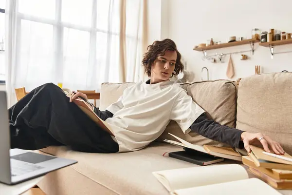 Young man relaxes on a couch at home, immersed in books and a cozy atmosphere during the day. — Stock Photo